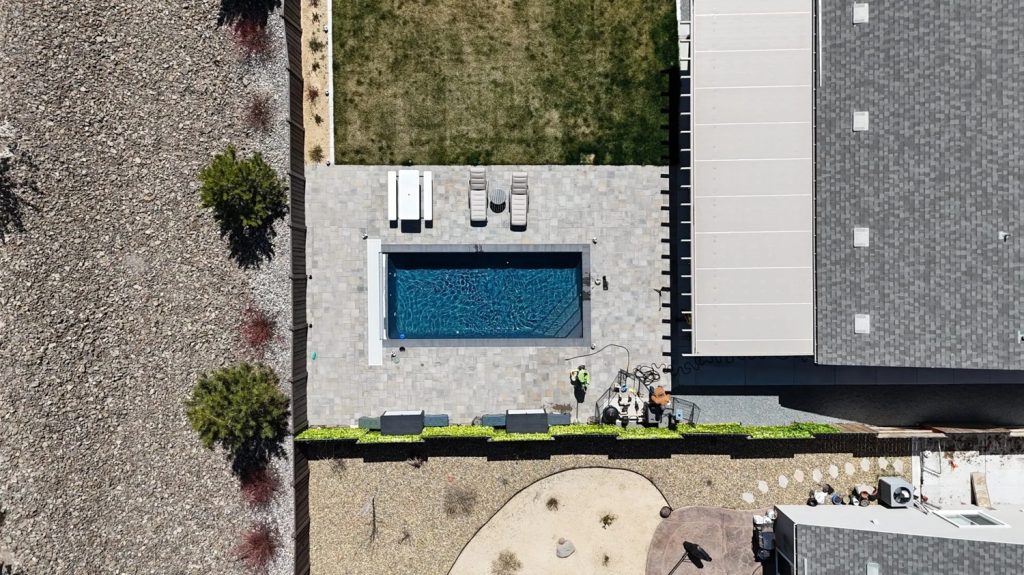 This aerial image shows a modern rectangular fiberglass inground pool set within a clean, structured backyard design. The pool is surrounded by a spacious gray paver patio, offering plenty of room for sun loungers, a table, and chairs—ideal for outdoor relaxation and entertaining.