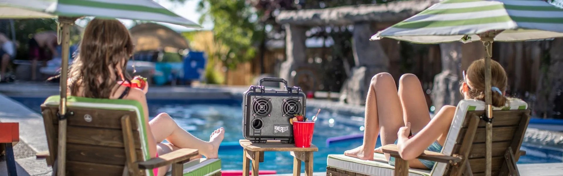 A vibrant backyard poolside scene on a sunny day. Two girls are relaxing in striped lounge chairs under green-and-white umbrellas, enjoying drinks and strawberries while facing a swimming pool.