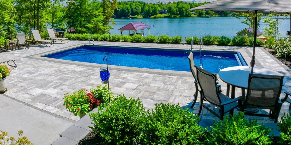 A beautiful fiberglass pool from Latham with a stone patio overlooks a scenic lake, surrounded by lounge chairs, patio seating, and lush greenery.