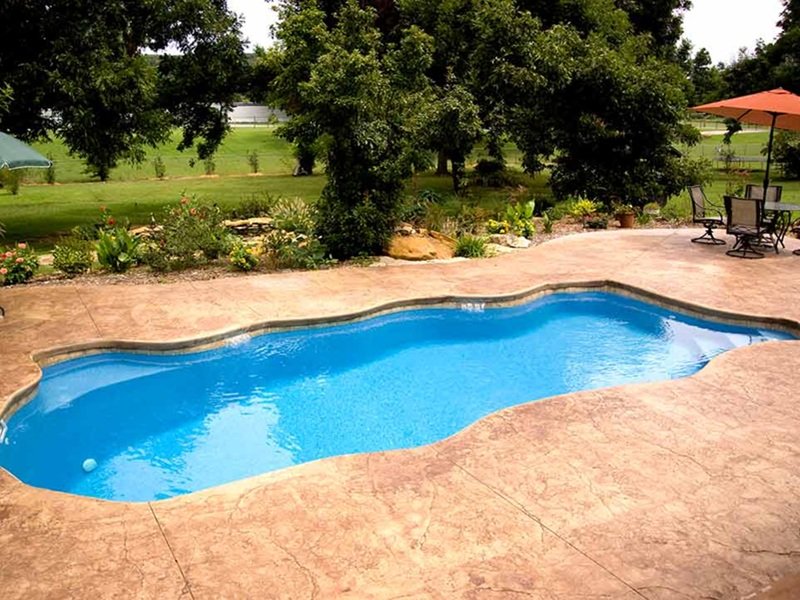A curvy inground pool with a stamped concrete patio is surrounded by lush greenery, trees, and a shaded seating area with a table and umbrella.