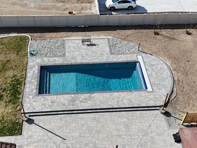 An aerial view of a backyard featuring a rectangular inground pool in Reno surrounded by stone pavers, with sections of grass, bare soil, and a hot tub area nearby.