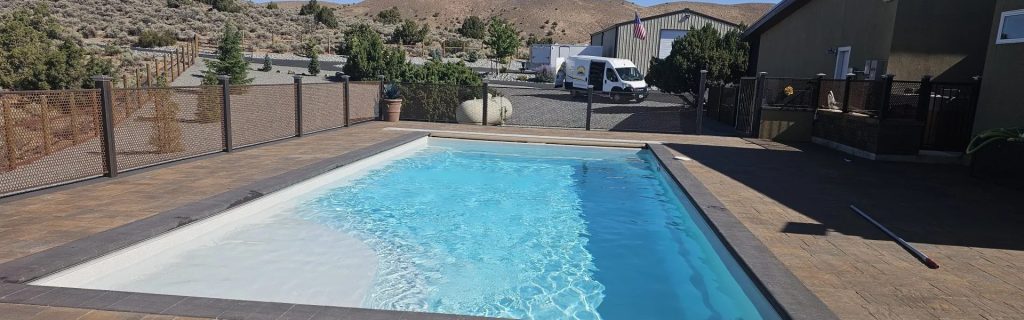 A rectangular in-ground swimming pool with light blue water, surrounded by stamped concrete decking, in a dry, hilly Northern Nevada landscape with nearby fencing and buildings.