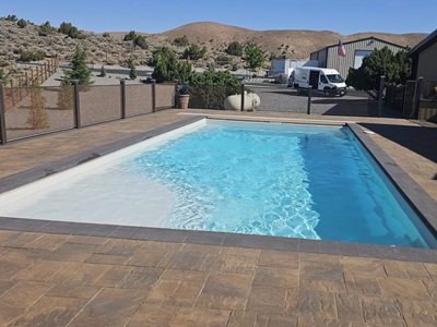 A rectangular in-ground swimming pool with light blue water, surrounded by stamped concrete decking, in a dry, hilly Northern Nevada landscape with nearby fencing and buildings.