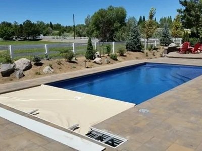 A rectangular inground pool with a partially extended automatic safety cover sits in a landscaped backyard with paver decking and seating areas.