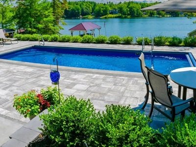 A beautiful fiberglass pool from Latham with a stone patio overlooks a scenic lake, surrounded by lounge chairs, patio seating, and lush greenery.