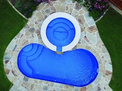 An aerial view shows a uniquely shaped inground pool with a connected circular spa, surrounded by a stone patio and landscaped greenery.