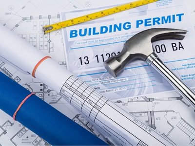 building permit papers with a hammer sitting on top, a ruler, and a rolled up piece of paper with a rubber band.