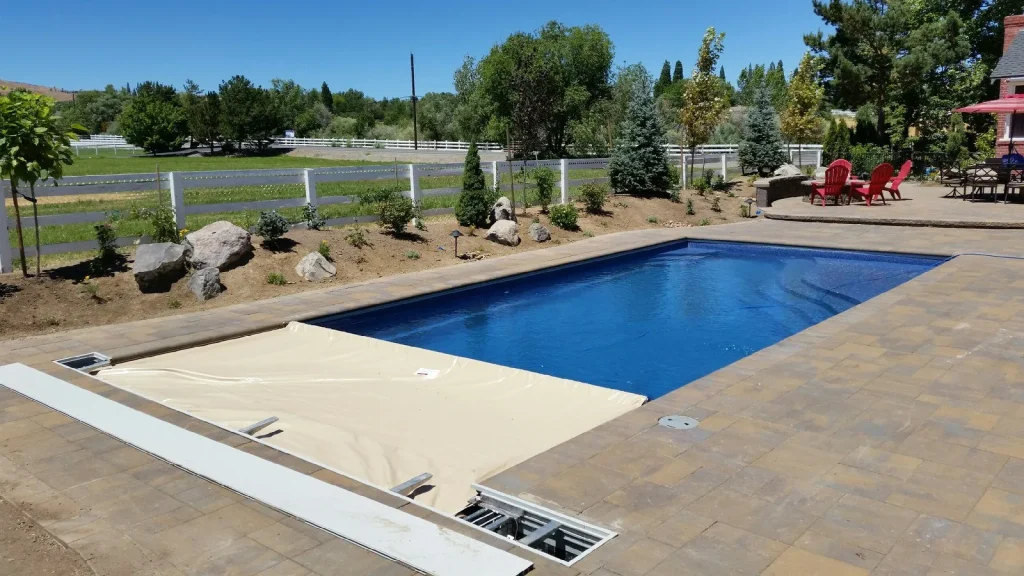 Automatic pool cover partially extended over a backyard swimming pool with a stone patio and landscaped yard.